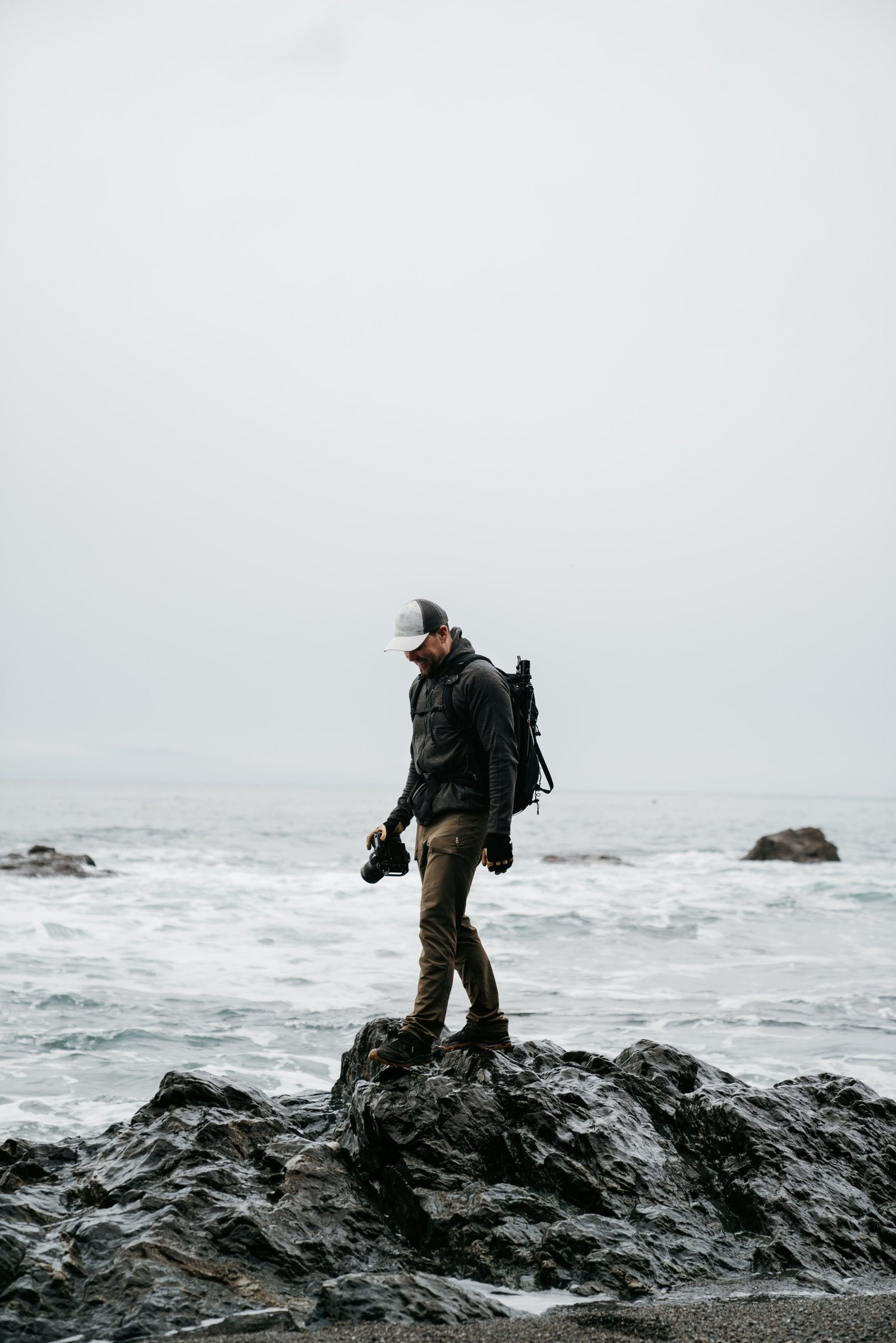Man standing on wet rocks at the beach, holding a camera, with ocean waves and rocks in the background under cloudy sky.